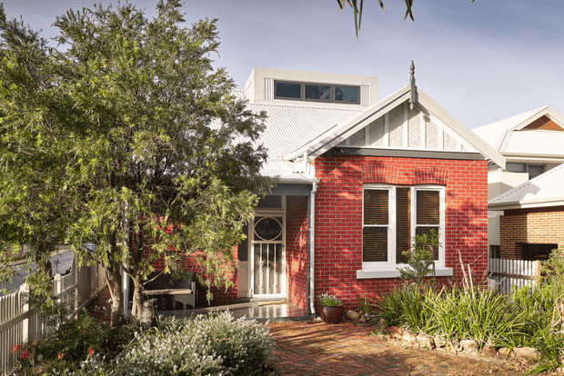 Street-facing view of a compact heritage-style brick home with a steep gabled roof, white painted detailing, decorative gable trim, narrow dormer-like clerestory window, and a small covered entry. The red brick facade is framed by mature garden planting and a brick-paved front path, with neighbouring homes visible at the sides.