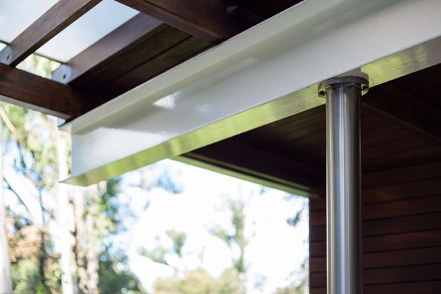 Close-up architectural detail of a covered outdoor structure, showing dark timber roof beams and soffit, a glossy white structural beam, and a cylindrical silver metal support column. Trees are visible beyond the open-sided structure.