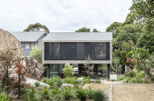 A contemporary two-storey residential extension set into a landscaped garden and rocky site. The dark, glazed upper level is screened with horizontal louvres, while the lower level has extensive glazing and a sheltered outdoor connection. A low-pitched metal roof, grey cladding, exposed rock, native-style planting, and a gravel or paved foreground are visible.