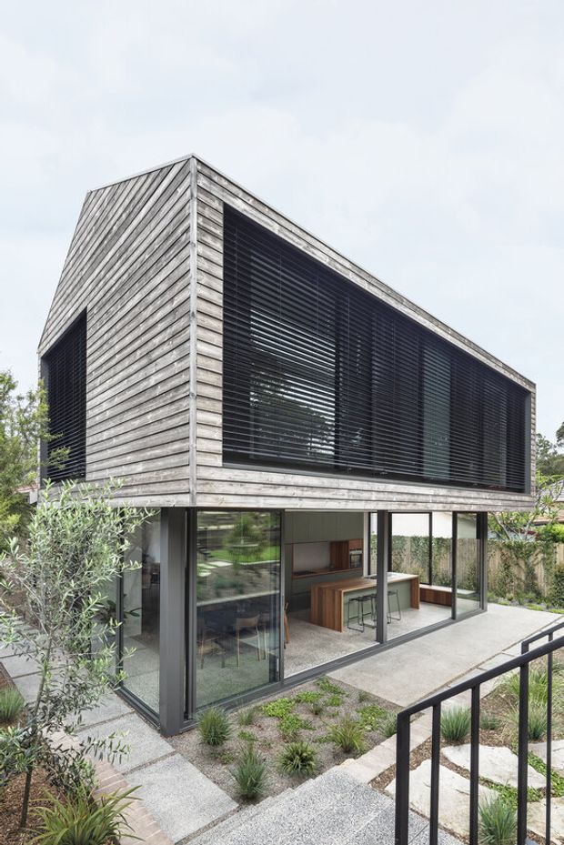 A compact contemporary two-storey house with a strongly geometric, box-like form. The upper level is clad in weathered horizontal timber boards and features a broad dark louvred opening, while the ground floor is largely glazed with dark-framed sliding doors and windows. A visible kitchen occupies the lower level, opening onto a concrete terrace and a landscaped garden with steps and black metal railings.