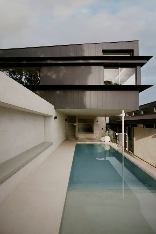 A contemporary multi-level residential courtyard viewed from beside a narrow rectangular swimming pool. The home features clean geometric volumes, dark horizontal roof and balcony projections, vertically ribbed grey-brown cladding, smooth light rendered walls, expansive glazing, and a pale paved pool surround. Built-in bench seating runs along the left wall, while white railings and a covered side structure frame the pool area.