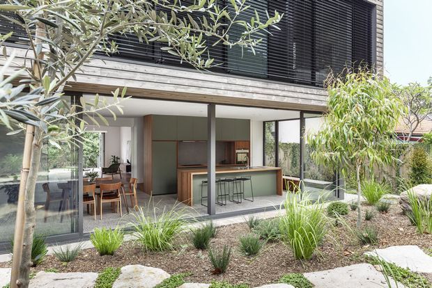 Contemporary two-storey residential extension viewed from a landscaped garden. The lower level has extensive glazing opening onto an open-plan kitchen and dining area with a timber island, pale green cabinetry, and bar stools. The upper facade combines horizontal timber cladding with dark external louvres or blinds. Naturalistic planting, gravel mulch, stone stepping slabs, and mature trees frame the foreground.