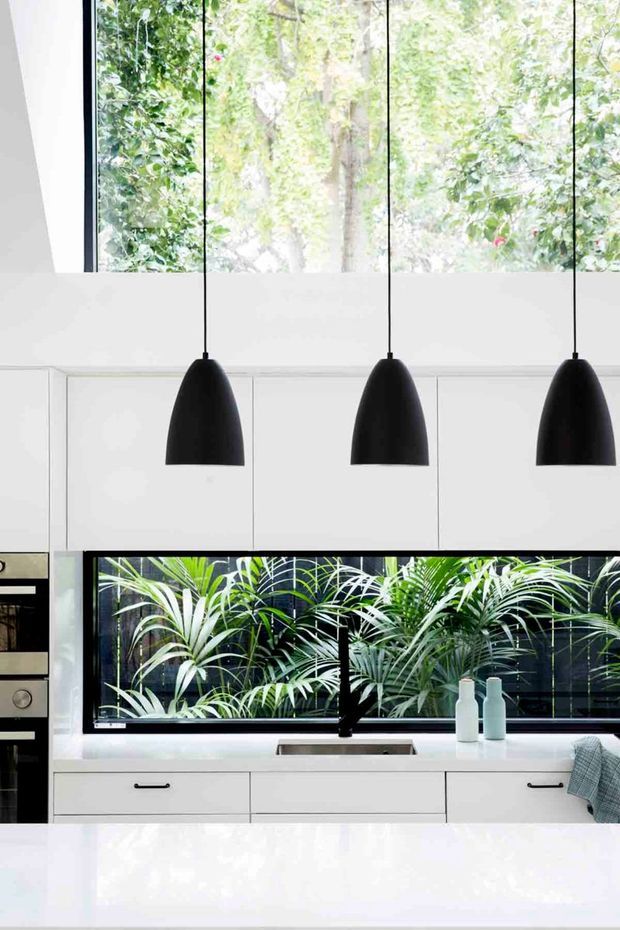 Bright contemporary kitchen with extensive white flat-panel cabinetry, a large white island, black-framed windows overlooking dense greenery, and three black pendant lights above the island. Integrated appliances and a sink beneath the garden-facing window complete the clean, high-contrast composition.