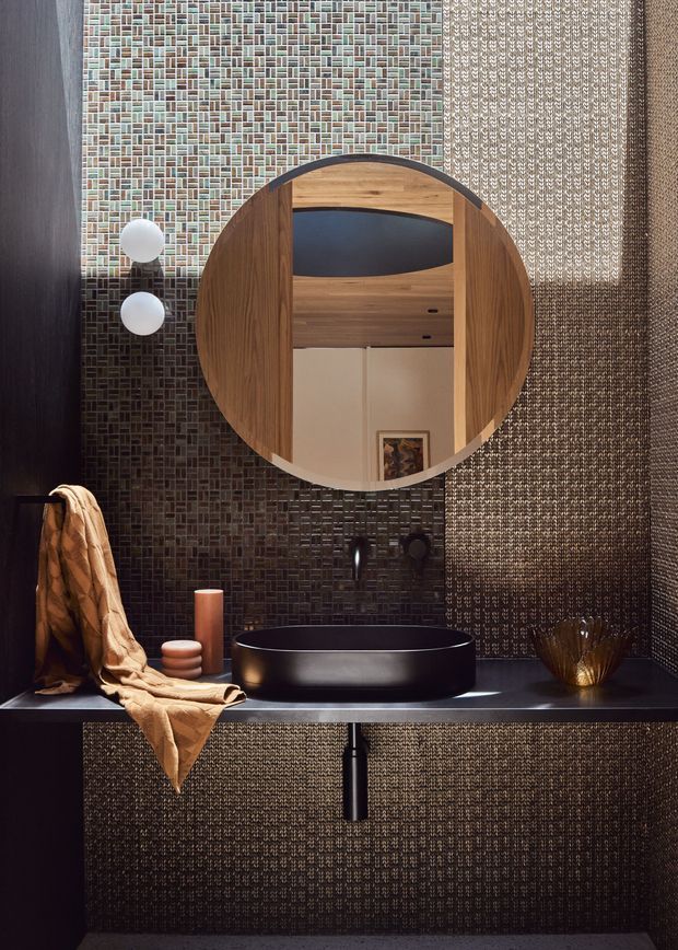 A compact, dramatically styled bathroom vanity featuring a matte black vessel basin on a dark countertop, black wall-mounted tapware, and a large round timber-framed mirror. The walls and vanity front are clad in dark small-format textured mosaic tile, with a contrasting woven-texture wall panel on the right. Warm timber, bronze-toned accessories, and soft amber lighting create a luxurious, moody atmosphere.