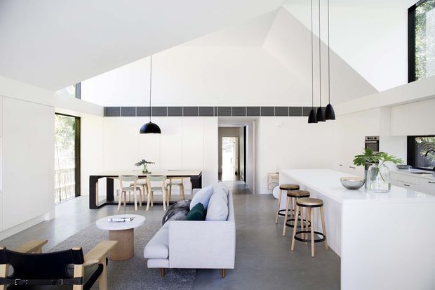 Bright open-plan contemporary interior combining a kitchen, dining area and living room beneath a dramatic pitched ceiling. White built-in cabinetry and walls contrast with a large white island, grey polished-looking floor, pale upholstered sofa, timber dining furniture and black pendant lights. Large windows provide abundant natural light and views of greenery.