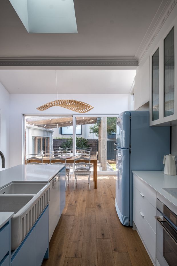 Bright, narrow contemporary kitchen viewed toward a glazed dining area and outdoor courtyard. The room features pale blue cabinetry, white benchtops, a matching retro-style blue refrigerator, timber-look flooring, white walls and ceiling, glass-front upper cabinets, and a sculptural woven pendant light. Decorative ceiling cornices and a large skylight add traditional detailing and natural light.