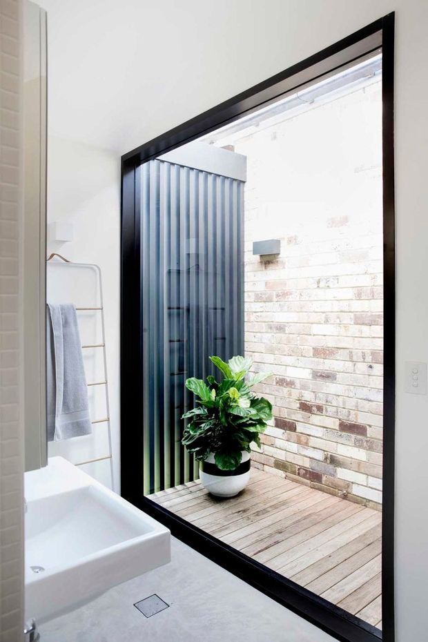 Bright contemporary bathroom featuring a white wall-mounted vanity and basin, pale grey floor, heated towel rail, and a large black-framed opening to a small outdoor lightwell. The lightwell has timber decking, an exposed pale brick wall, and a dark vertical slatted privacy screen, with a potted leafy plant in the foreground.