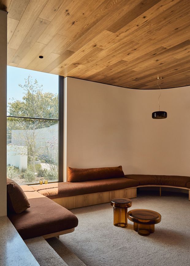 Warm, refined living room with a low built-in curved sectional sofa wrapping around the space, paired with amber-toned glass coffee tables. Full-height glazing opens to a planted exterior, while the ceiling is clad in richly grained timber boards and the walls are softly neutral. A suspended dark pendant light provides a simple focal point.