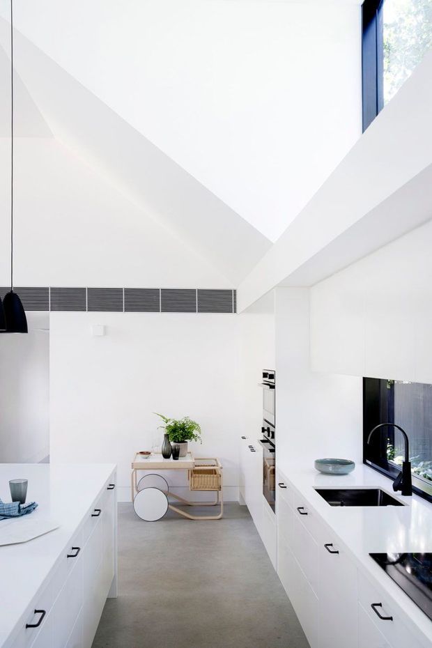 Bright, minimalist white kitchen with parallel banks of cabinetry, a central walkway, integrated appliances, black hardware and fixtures, and a concrete-look floor. A large angular ceiling volume creates a dramatic architectural feature, with high-level glazing bringing in natural light. A small dining or serving area is visible at the far end.