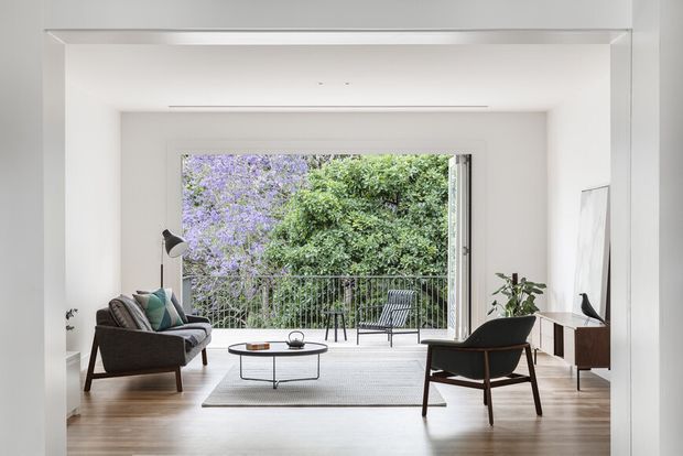 Bright, minimalist living room with pale timber flooring, white walls and ceiling, and a large glazed opening onto a balcony or terrace. The room contains a dark upholstered sofa, matching lounge chair, low coffee table, floor lamp, sideboard, and potted plant. Lush greenery and flowering trees are visible outside, creating an airy indoor-outdoor connection.