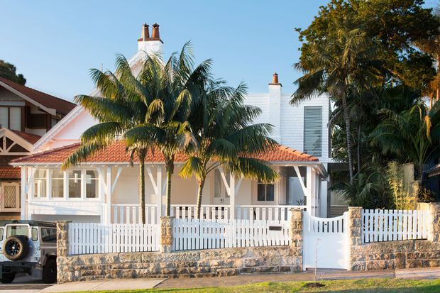 A bright exterior view of a low-rise residential house with white weatherboard walls, decorative gable detailing, a terracotta tiled pitched roof, prominent chimneys, a covered front veranda, and white picket fencing. Tropical palms and mature vegetation frame the street-facing facade, while a stone retaining wall forms the front boundary.
