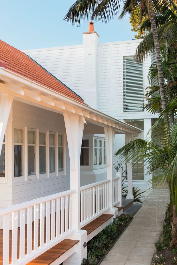 Exterior view of a bright coastal-style residence, showing a covered veranda with white railings and posts, timber decking, white weatherboard walls, red tiled pitched roof, and a taller adjoining white brick-clad volume with a chimney and louvred window. Palm trees and a narrow paved walkway frame the facade.