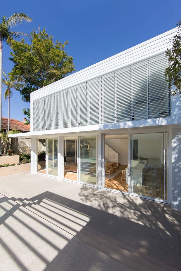 Bright exterior view of a contemporary two-storey residential extension with a white rendered facade and a prominent upper-level screen of horizontal louvres. Full-height glazed sliding doors open onto a broad concrete terrace, revealing a light-filled dining and living area inside. Strong linear shadows from the louvres fall across the terrace, with mature tropical and subtropical planting around the property.