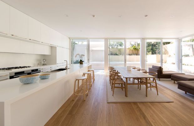 Bright, open-plan contemporary kitchen and dining area with extensive white built-in cabinetry, a large white island with integrated cooking appliances and seating, and a timber dining table surrounded by chairs. Full-height glazing opens the room visually to a planted courtyard or outdoor area. Warm timber flooring, neutral furnishings, and abundant daylight create an airy, refined interior.
