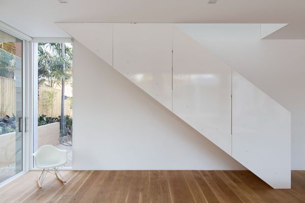 Minimalist interior with a sculptural white built-in storage or stair enclosure forming a strong diagonal profile across the wall and ceiling. The room has warm timber flooring, extensive floor-to-ceiling glazing on the left, and a white molded chair. Outdoor planting and neighbouring structures are visible through the glazing.