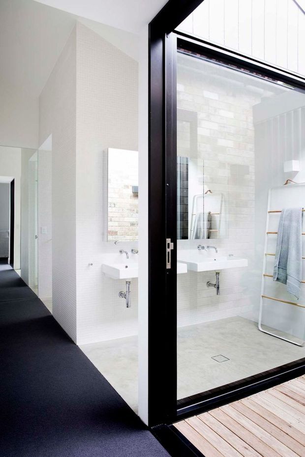 Bright minimalist bathroom viewed through a black-framed glazed door, with white textured wall tiling, a wall-mounted white basin, exposed plumbing, pale floor tiles, a frameless mirror, and a freestanding ladder-style towel rail. A dark carpeted hallway and timber decking are visible around the bathroom entrance.