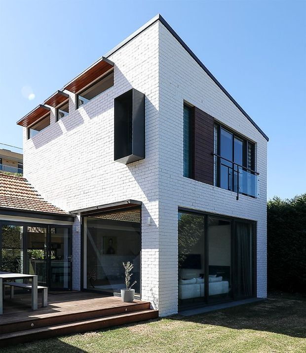 Exterior view of a contemporary two-storey residential extension or townhouse-like dwelling. The building has a sharply pitched gable roof, white-painted brick walls, dark metal-framed glazing, projecting upper-level window awnings with timber soffits, and a timber deck adjoining a glazed living area. A tiled-roof lower wing and landscaped lawn are also visible.