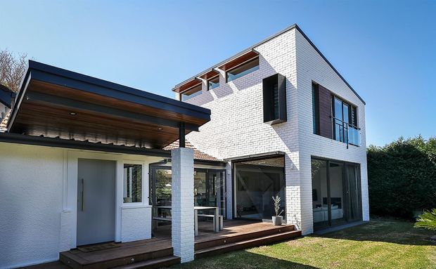 Exterior view of a contemporary two-storey residence with white brick walls, dark-framed glazing, projecting box forms, and a broad timber-lined covered patio or carport structure. The home includes a timber deck, outdoor seating area, and landscaped lawn, with a clear blue sky and mature hedging around the property.