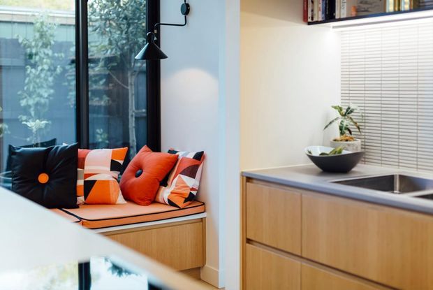 A compact contemporary kitchen and built-in window-seat nook. Warm timber cabinetry runs beneath a dark benchtop, with a stainless-steel sink beneath a partially lowered blind. The adjacent window seat has a peach cushion and bold orange, red, black and white cushions, framed by large black window surrounds. A black wall-mounted pendant light and open shelving add contrast.
