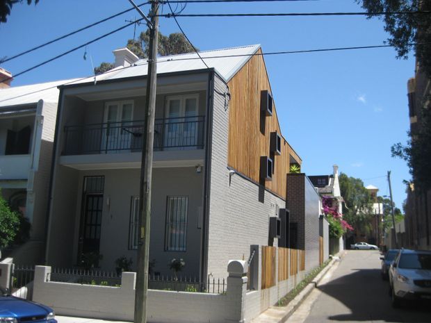 Street-level view of a narrow urban residential lane lined with attached multi-storey homes. The foreground house has a light grey rendered or masonry facade, barred ground-floor windows, a recessed balcony with black metal balustrade, and a pitched corrugated metal roof. A newer adjoining volume features warm vertical timber cladding and dark-framed windows, creating a contemporary townhouse-style composition. Utility poles, overhead wires, paving, and neighbouring residences are visible.