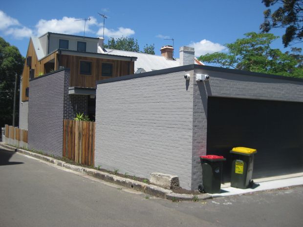 Exterior street view of a compact multi-level residential building with a prominent grey brick garage, timber-clad upper storey, pitched metal roof sections, and a narrow timber privacy fence. The image is taken from the street, with the garage and facade dominating the frame.