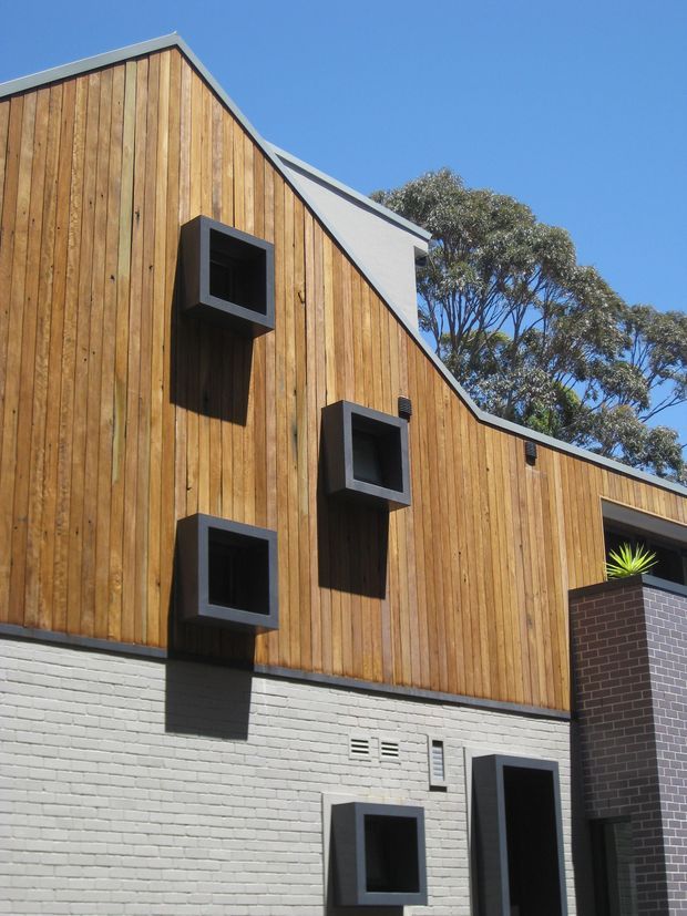 Exterior view of a contemporary multi-level building with a steep gabled roof form, warm vertical timber cladding, and a contrasting painted white brick lower facade. Several projecting dark-framed square windows punctuate the facade, with a dark brick entry volume at the right. Clear blue sky and mature trees form the background.