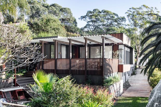 A compact single-storey dwelling nestled in lush landscaped greenery, with dark timber or metal cladding, extensive glazing, covered verandah-like decks, corrugated roof planes, and a timber walkway approaching the entrance. The home has a contemporary pavilion-like form integrated into a leafy garden setting.