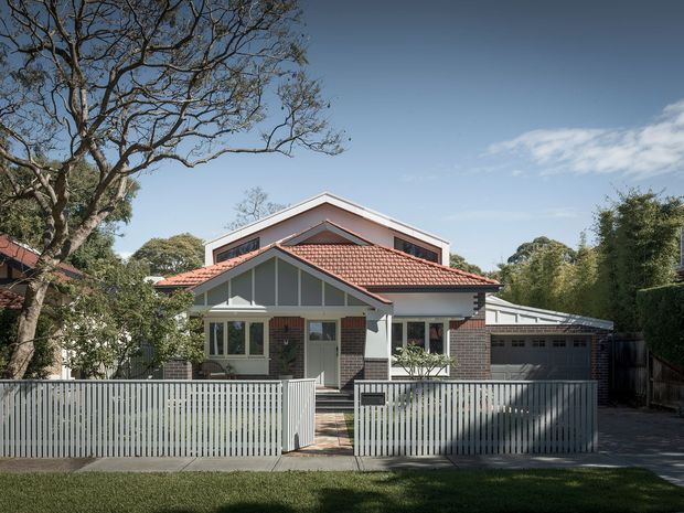 Street-facing view of a traditional suburban house with a red terracotta tiled gabled roof, exposed brick walls, white timber detailing, and a white vertical-slat front fence. A front porch, entry path, and attached single-car garage are visible, with mature trees surrounding the property.