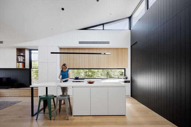 Bright contemporary kitchen in an open-plan interior, featuring a large white island with integrated cabinetry, pale timber flooring, white walls and cabinetry, a timber overhead cabinet, black linear pendant lighting, and a dramatic dark vertically panelled feature wall. High-level glazing and clerestory windows bring in abundant natural light. A small dining/work table and stools sit beside the island, with a living area partially visible beyond.