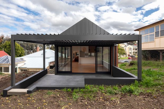 Exterior view of a compact contemporary house extension or pavilion on a sloping suburban site. The dark charcoal structure has a pronounced triangular gable roof form, projecting covered deck or carport-like frame, broad black-framed glazed doors, and a warm timber-floored interior with a visible white kitchen beyond. Neighbouring houses and landscaped terrain surround the building.