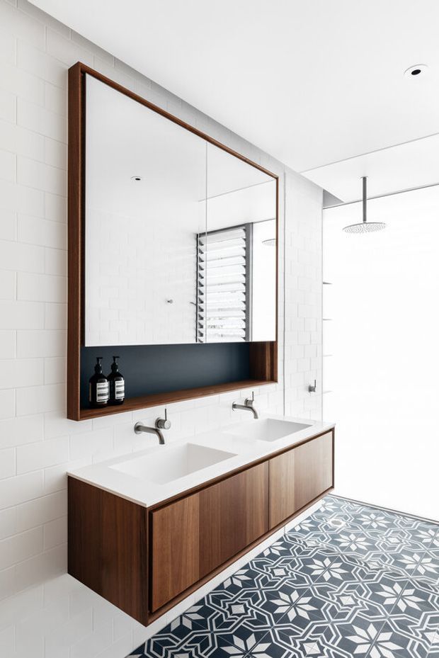 Bright contemporary bathroom featuring a wall-mounted double vanity with dark timber cabinetry, white integrated basins, brushed metal wall-mounted taps, and a large timber-framed mirrored cabinet with an open dark recessed shelf. The walls are clad in white rectangular tiles, while the floor has bold patterned geometric tiles. A walk-in shower with a ceiling-mounted rain shower is visible at the right.
