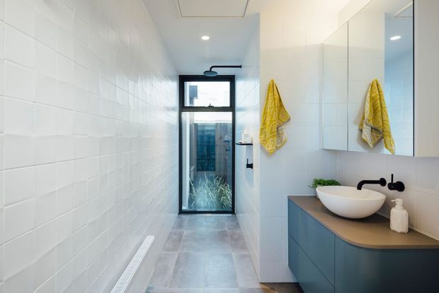 A narrow, contemporary bathroom with white tiled walls, grey tiled flooring, a floating dark blue vanity with a rounded stone benchtop, white vessel basin, matte black wall-mounted tapware, mirrored cabinetry, and a glass door opening to a small planted outdoor area. A walk-in shower is positioned toward the rear, with a skylight and recessed ceiling lighting providing bright illumination.