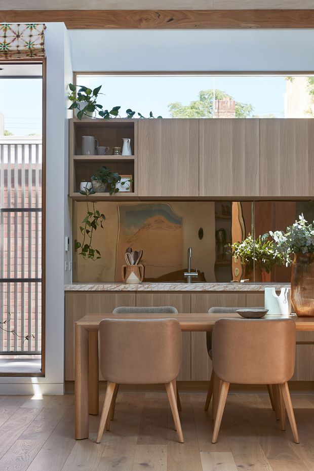 A bright, compact open-plan kitchen and dining area with warm timber cabinetry, a marble-patterned benchtop, reflective metallic splashback, and a timber dining table with upholstered chairs. The kitchen features open shelving, a long horizontal window, indoor plants, and abundant natural light. A glazed balcony or exterior walkway is visible at left.