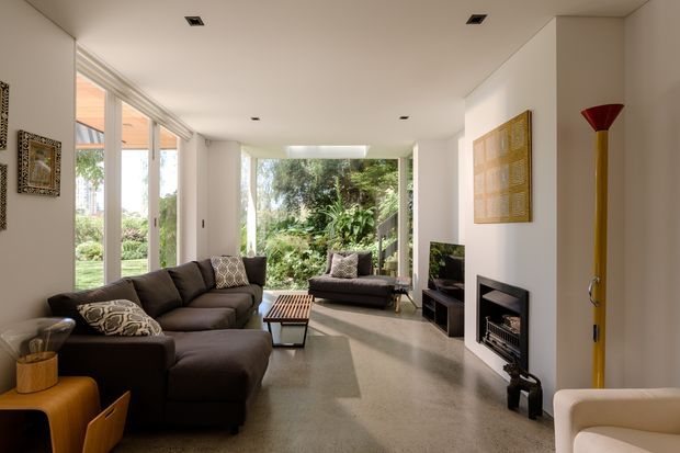 Bright contemporary living room with a large dark sectional sofa, additional seating, a low coffee table, television, and compact fireplace. Full-height glazing along the side and at the rear opens views toward a lush garden, while white walls, a polished light-toned floor, recessed ceiling lights, and curated artwork create a clean, refined interior.