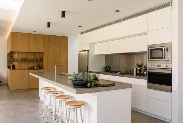 A bright, spacious contemporary kitchen with a large central island and four stools. The kitchen features white full-height cabinetry, integrated stainless-steel appliances, a timber wall with open shelving and concealed storage, a neutral stone island benchtop, and a linear cooktop splashback. Recessed lighting and a long linear air-conditioning grille contribute to the clean modern appearance.