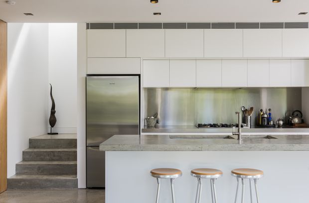 Bright contemporary kitchen with a long island and three stools, white floor-to-ceiling cabinetry, stainless-steel refrigerator and splashback, and a pale stone benchtop. A short set of concrete or stone steps leads to an adjacent area at left. Recessed ceiling lighting and linear air-conditioning vents contribute to the clean, minimal composition.