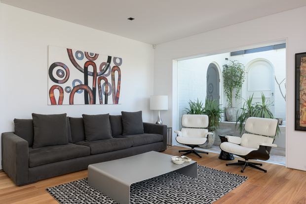 A bright contemporary living room with a large charcoal sectional sofa, two cream lounge chairs, a low rectangular grey coffee table, and a black-and-white geometric rug. White walls and ceiling contrast with warm timber flooring. A large glazed opening looks onto a planted courtyard with decorative arched architectural details, while abstract artwork and a floor lamp add visual interest.