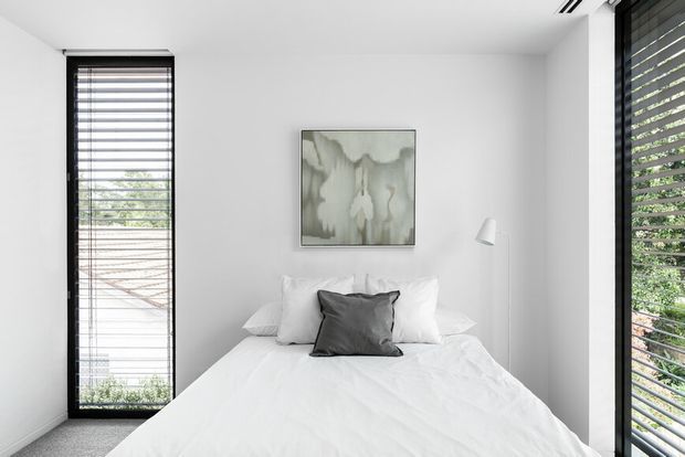 A compact, bright bedroom with a neatly made white bed positioned between two tall windows. The room has white walls and ceiling, dark window frames with horizontal blinds, a small white wall-mounted reading light, and a muted abstract artwork above the bed. The restrained monochromatic palette creates a clean, contemporary atmosphere.