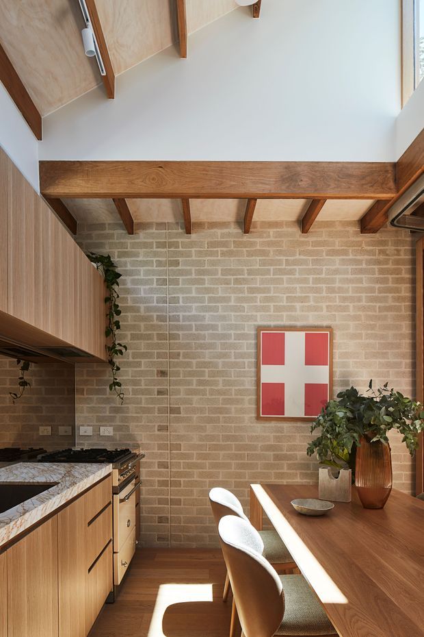 A warm, compact kitchen-dining space with light timber cabinetry, a dark stone benchtop, exposed timber ceiling beams, and a pale brick feature wall. A timber dining table with upholstered chairs occupies the right side, while indoor plants and simple framed artwork add visual interest. Strong natural light enters from the upper right, creating a bright, inviting atmosphere.