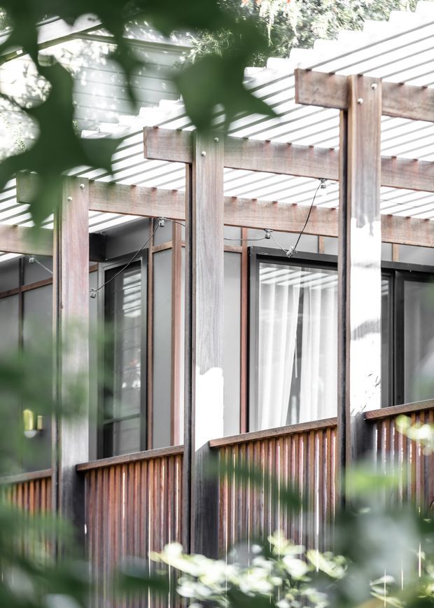 A close-up exterior view of a contemporary house or deck structure, framed through soft-focus foliage. The facade features vertical timber posts and balustrading, dark-framed glazed doors or windows, a translucent or slatted overhead pergola, and exposed timber beams with visible exterior string lighting. The composition has a warm natural-material palette and strong filtered daylight.