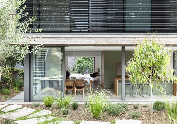 Exterior view of a contemporary multi-level home, focused on a glazed ground-floor living and dining area opening onto a landscaped garden. The facade combines dark horizontal window blinds, large glass sliding doors, and a light timber-clad band, with extensive low-maintenance planting and stepping-stone paving in the foreground.