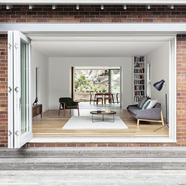 A bright, refined living room viewed from a wide open exterior opening, with white walls, pale timber flooring, a grey sofa, green lounge chair, low black coffee table, and built-in bookcase. The room extends to a dining area with large glazing overlooking a rocky landscaped setting. The exterior is clad in exposed reddish-brown brick with broad timber decking and white-framed bi-fold doors.