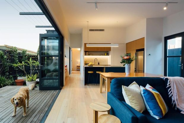 Open-plan contemporary home interior with a kitchen, dining area and living room opening onto a timber deck and planted outdoor courtyard. The space features pale timber flooring, white walls, warm timber cabinetry, a dark kitchen island, expansive black-framed glazing and a deep blue sofa with colourful cushions.