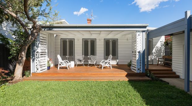 A bright single-storey residential courtyard-style exterior with a covered timber deck/verandah. The facade has pale weatherboard cladding, white-framed barred windows, decorative louvred or slatted side screens, and a low-pitched roof. White outdoor seating and a small table occupy the deck, which opens onto a lawn and connects to a side wing by timber steps.