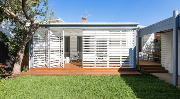 Bright exterior view of a single-storey house with a white weatherboard facade and an expansive timber deck enclosed by white horizontal slatted privacy screens. Steps connect the deck to a side entry, while a lawn and mature tree occupy the foreground. The design combines a low-pitched roof, screened outdoor living space, and a contemporary extension or adjoining volume.