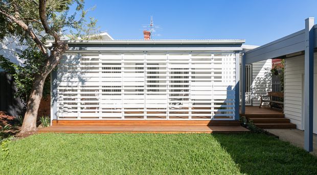 Exterior view of a low-rise residential extension with a broad white horizontal slatted screen enclosing a glazed living area. A timber deck and steps connect to a side entry, while a rendered or painted horizontal-clad wing frames the right side. The scene includes a front lawn and mature tree under bright blue sky.