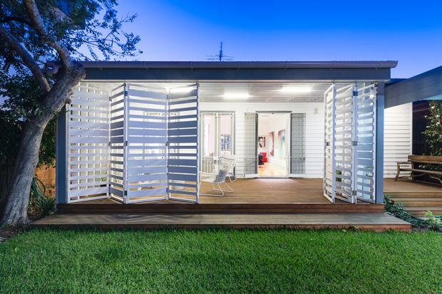 A compact contemporary pavilion-style dwelling viewed from the lawn at dusk. The single-storey structure has a flat roof, white horizontal cladding, extensive white louvred screening around a timber deck, and illuminated glazed openings leading into the interior. A mature tree frames the left side, while a covered outdoor seating area extends to the right.