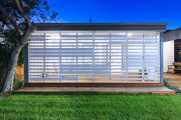 Exterior view of a contemporary single-storey garden structure or enclosed outdoor room, featuring extensive white horizontal louvred screening over glazing, a shallow flat roof, and a timber deck. A lawn and mature tree sit in the foreground, with a timber-clad building and outdoor seating visible to the right.