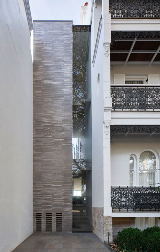 Narrow urban architectural view showing a contemporary infill facade inserted beside an ornate historic terrace building. The new element has a tall vertically stacked grey stone-tile wall, a narrow full-height glazed opening, and a dark paved entry court, contrasting with the adjacent white rendered facade, arched window, and elaborate black decorative metal balconies.