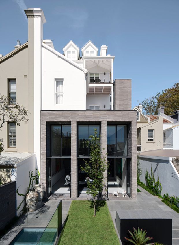 A vertically framed view of a narrow multi-storey urban residence showing a contemporary rear extension attached to a pale traditional house. The extension has a rectangular grey masonry-clad facade with large black-framed floor-to-ceiling glazed openings, opening onto a small landscaped courtyard with lawn, paving and a plunge pool. Above, the original house features white rendered walls, pitched gables, dormer windows, chimneys and a covered balcony. The composition contrasts heritage-style residential architecture with a modern addition under a clear blue sky.